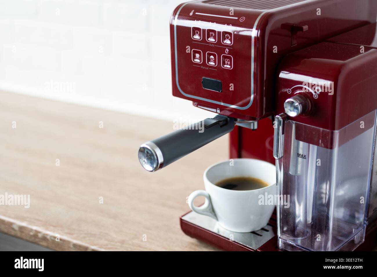 Primo piano di una macchina per caffè espresso rossa che prepara caffè fresco in una tazza bianca su un ripiano in legno in una cucina moderna. Caffè casalingo minimalista Foto Stock