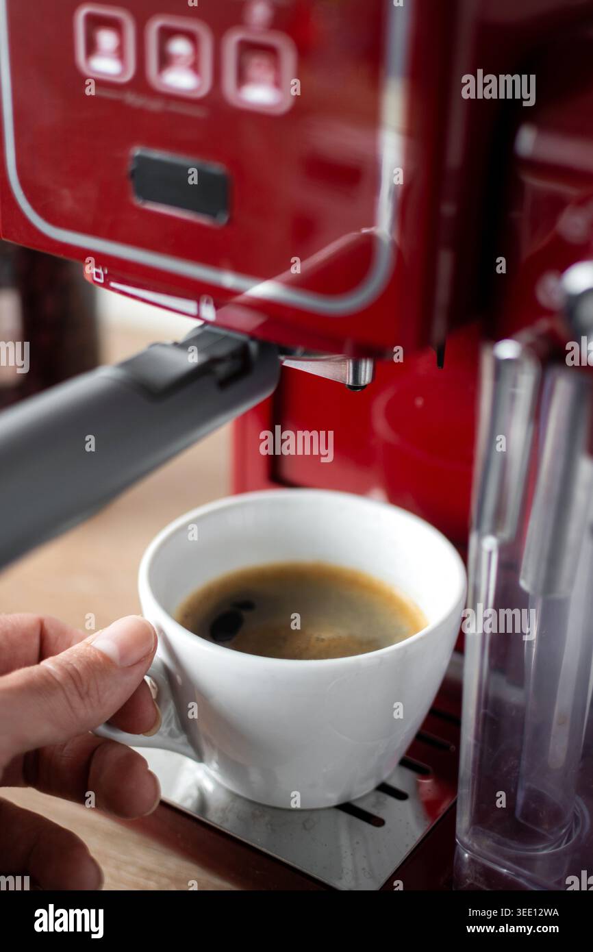 Primo piano di una persona che tiene in mano una tazza bianca riempita di caffè espresso fresco da una macchina da caffè rossa in una cucina moderna. Preparati al caffè casalingo Foto Stock