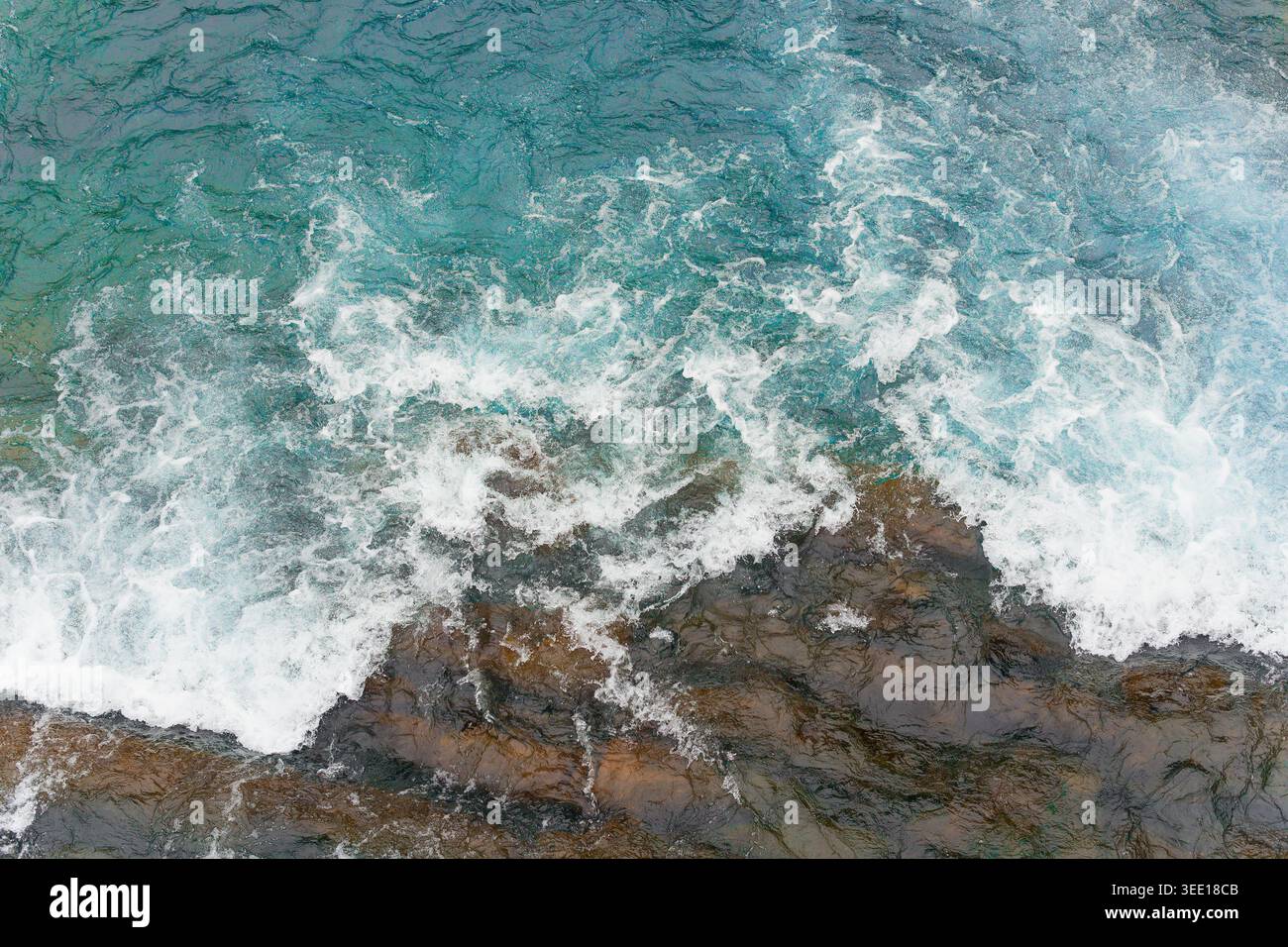 Primo piano dall'alto di un'aspra costa rocciosa con spruzzi d'acqua turchese dinamica, schiuma marina bianca che gira contro le pietre intemprate in vividi dettagli naturali Foto Stock