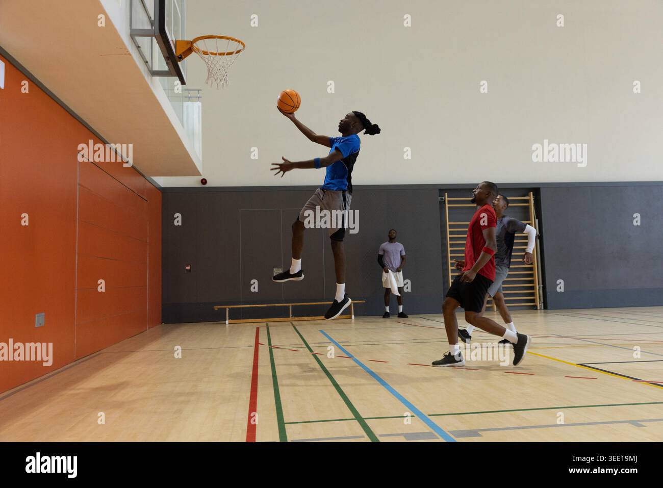 Diversi giocatori maschili di abbigliamento sportivo che giocano a basket in palestra, saltano a strapiombo verso il cerchio. Palla, campo, backboard, compagni di squadra, salto, perforazione dinamica Foto Stock