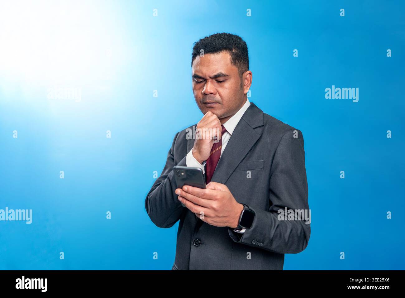 Ritratto di un dirigente indonesiano in tuta, tenendo uno smartphone con una mano sul mento e riflettendo profondamente su uno sfondo blu dello studio. Foto Stock
