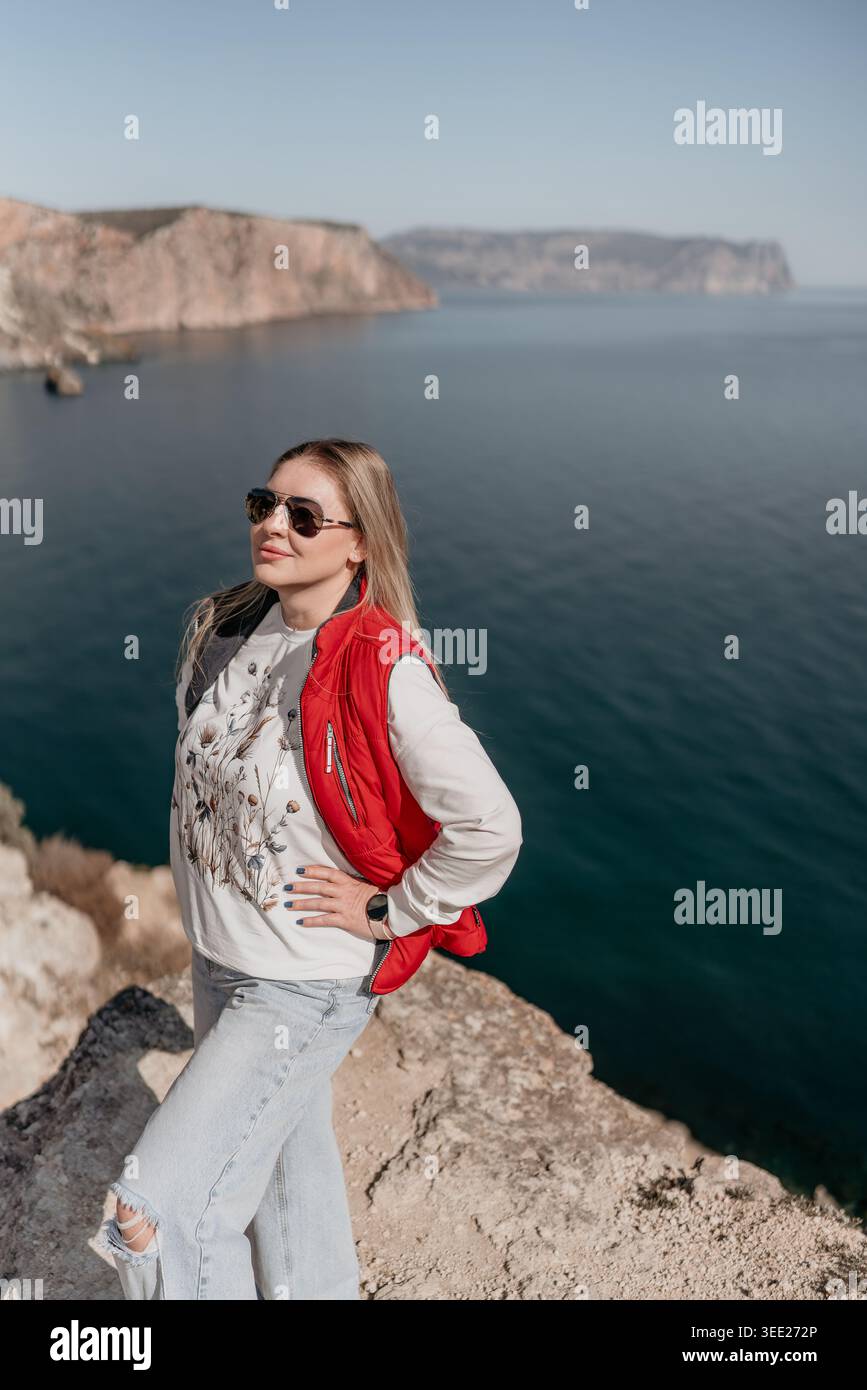Donna viaggiatrice sulla scogliera dell'oceano in un gilet rosso in piedi su un bordo roccioso che si affaccia sull'acqua Foto Stock