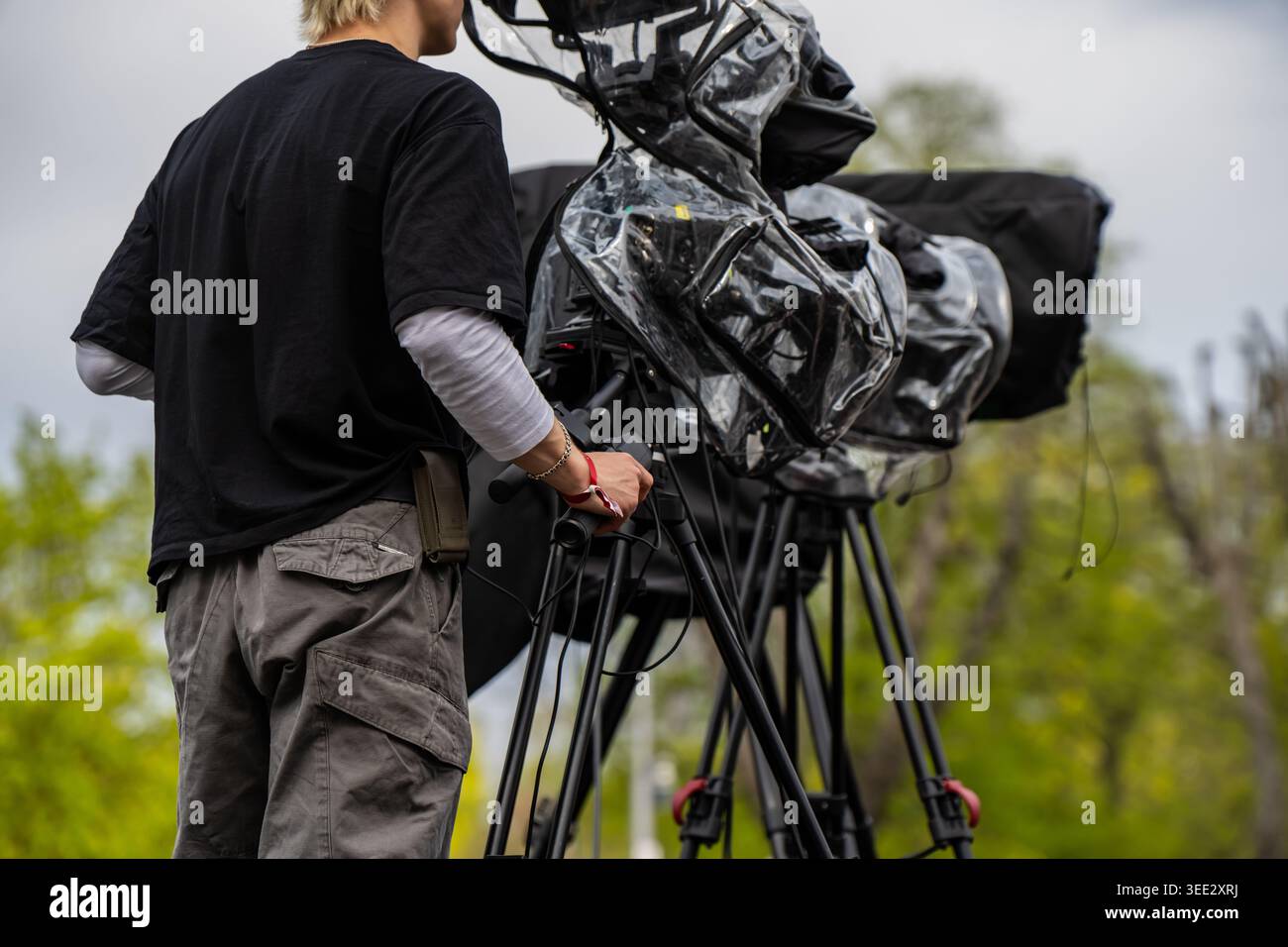 Operatore di telecamere professionale che gestisce apparecchiature di trasmissione di fascia alta con coperture antipioggia durante la produzione di eventi in diretta all'aperto in un parco cittadino per teleobiettivo Foto Stock