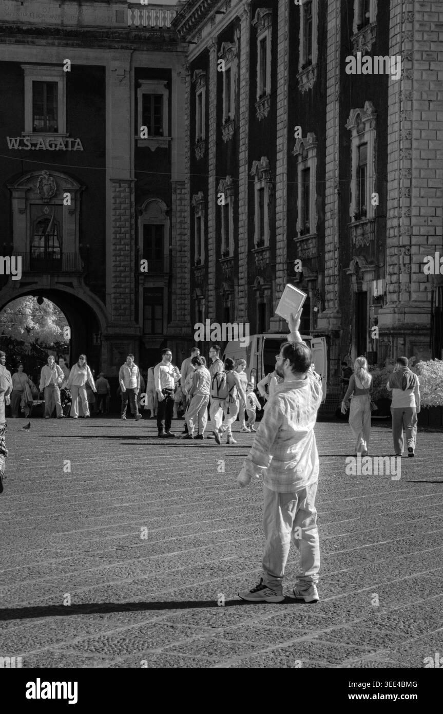 Un uomo si trova in una vivace piazza di Catania, Sicilia, Italia, tenendo un libro in alto con una mano. Il rendering della scena è in bianco e nero, captur Foto Stock