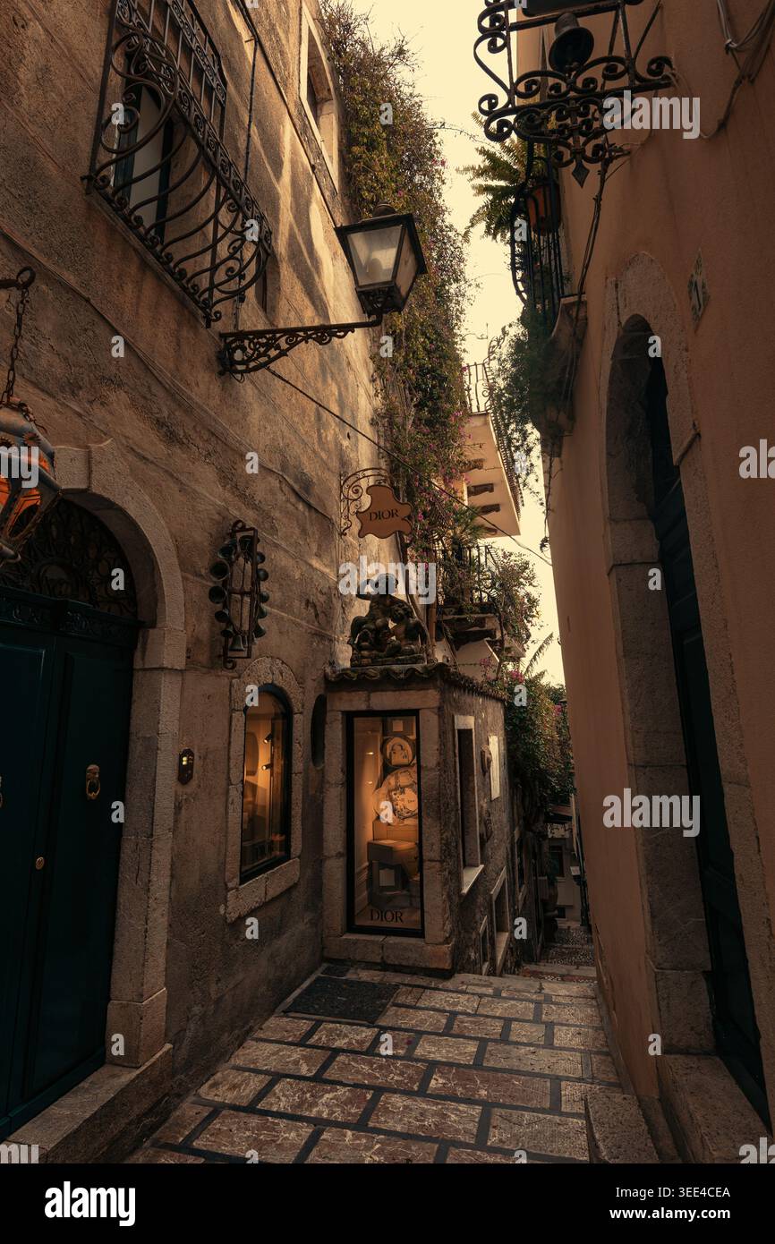 Una vista su uno stretto e antico vicolo in pietra a Taormina, Sicilia, caratterizzato da edifici storici. Un piccolo punto vendita Dior con elementi di visualizzazione illuminati Foto Stock