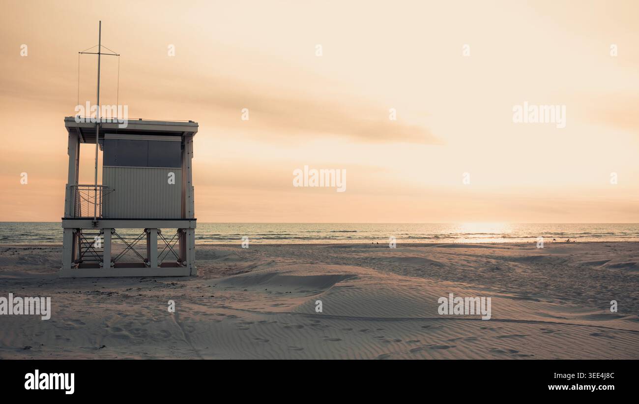 A guardia della solitaria torre del bagnino della costa sorge sulla spiaggia vuota del Baltico a Liepaja, incorniciata da un orizzonte aperto e dalla luce soffusa della sera. Solitudine e S. Foto Stock