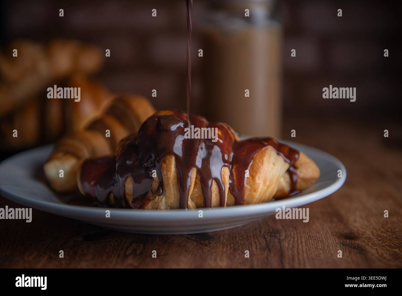 Pasticceria o panetteria, sfoglia ricoperta di cioccolato fondente, su un piatto bianco, con caffè freddo o frappe sullo sfondo Foto Stock