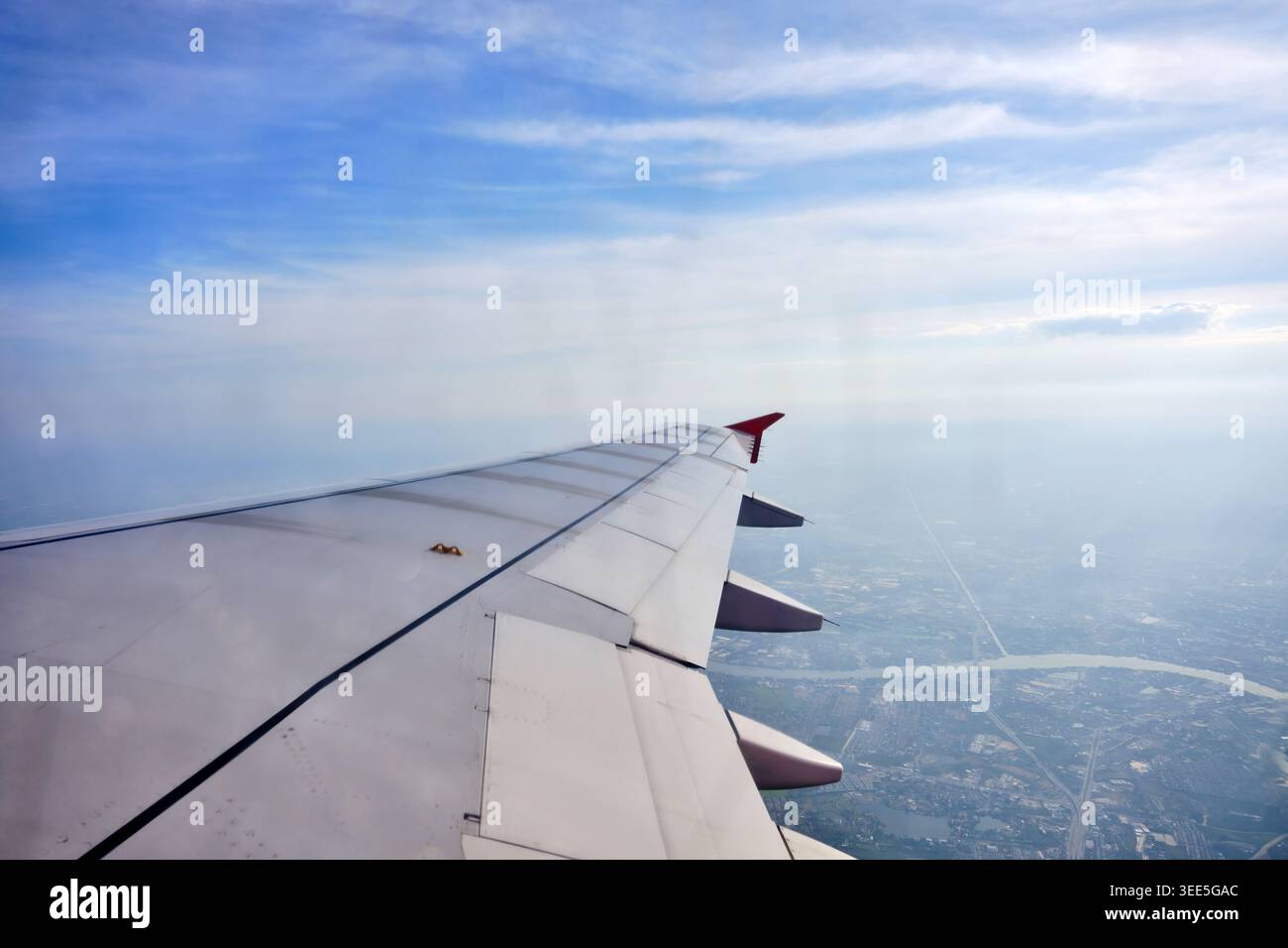 una vista dalla finestra dell'aereo che ha un'ala con aletta rossa nel cielo con nuvole e città sottostante. Foto Stock