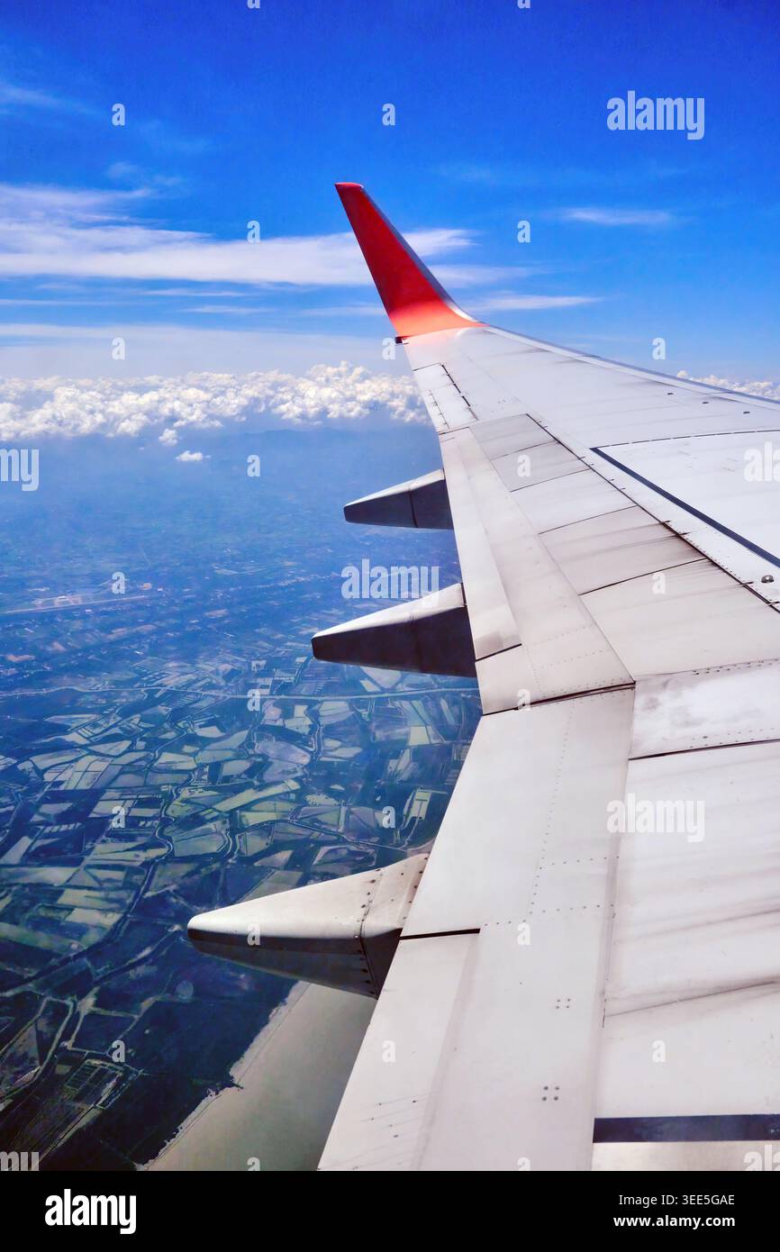 una vista dalla finestra dell'aereo che ha un'ala con aletta rossa nel cielo con nuvole e città sottostante. Foto Stock