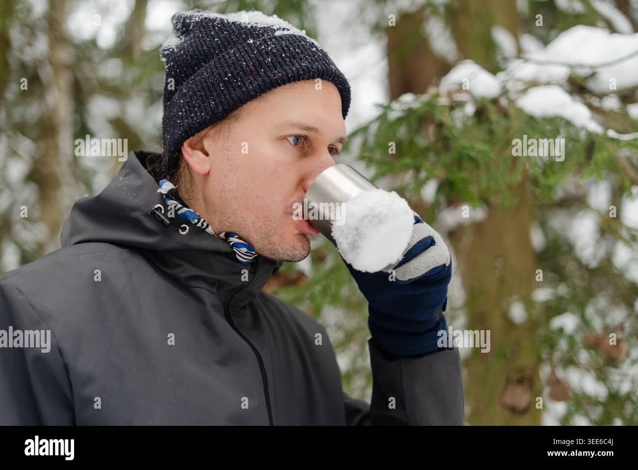 Uomo di mezza età sorseggiando tè caldo da un thermos d'argento in una foresta innevata. Un gentiluomo con una sciarpa bianca e blu a fantasia si gode una pausa con una bevanda calda durante una passeggiata con il freddo tra gli alberi innevati Foto Stock