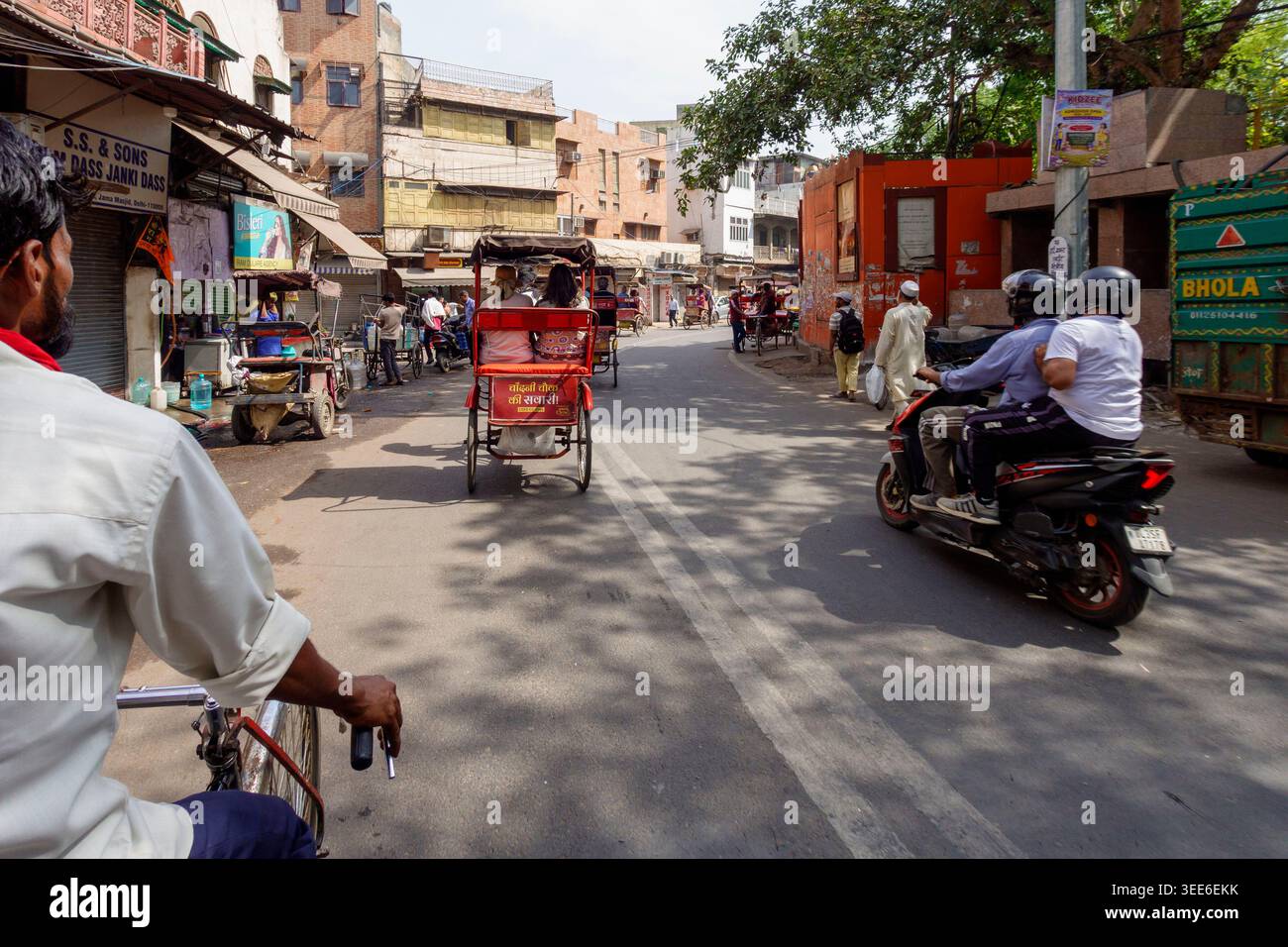 Vivace scena urbana della vecchia Delhi, India, con pendolari che viaggiano in bicicletta in risciò e moto attraverso una stretta strada della città costeggiata da negozi Foto Stock