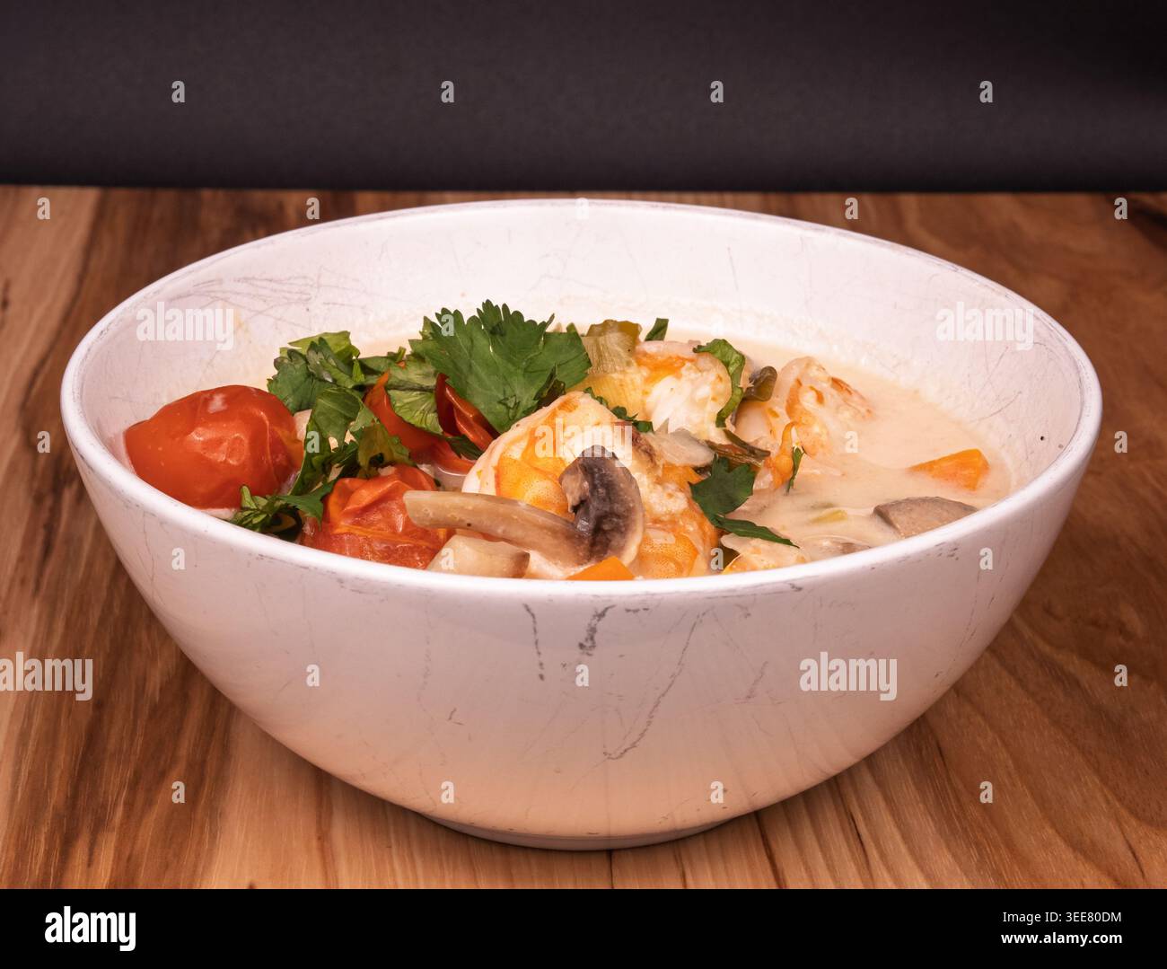 Foto a livello di occhi di una ciotola fumante di zuppa di gamberi tailandesi su uno sfondo scuro, che mette in risalto i colori vivaci dei frutti di mare e delle erbe aromatiche. Contromisure elevate Foto Stock