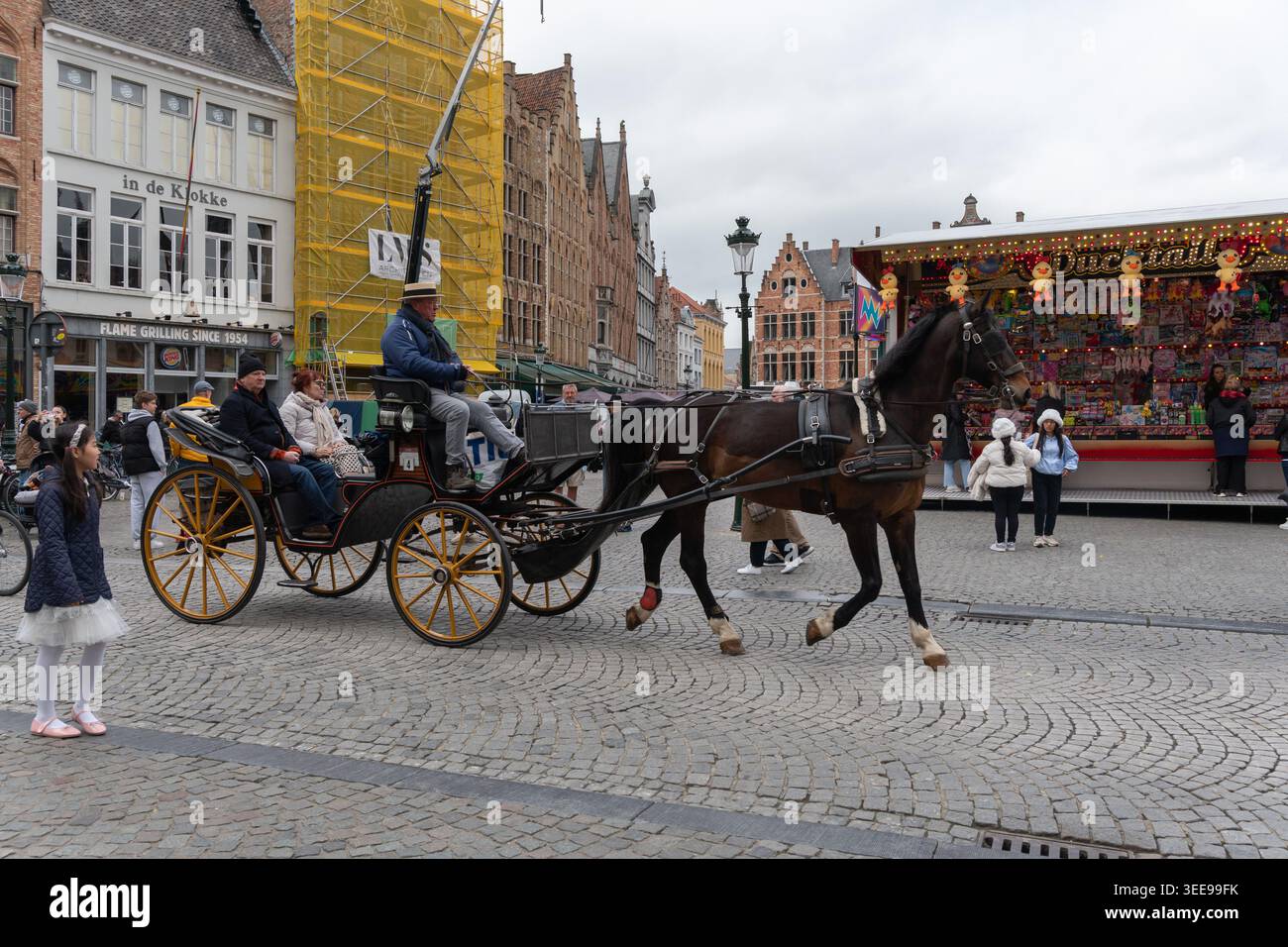 Bruges, Belgio - 3 aprile 2026: Grote Markt mostra una carrozza trainata da cavalli che attraversa la piazza acciottolata con autista, pedoni, chiosco alimentare, ponteggi Foto Stock