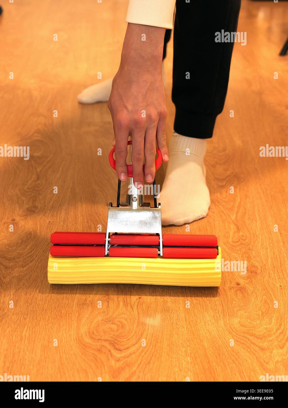 Persona che utilizza un panno in spugna giallo con rulli rossi per pulire un pavimento in legno in interni, che rappresenta lavori domestici e lavori domestici Foto Stock