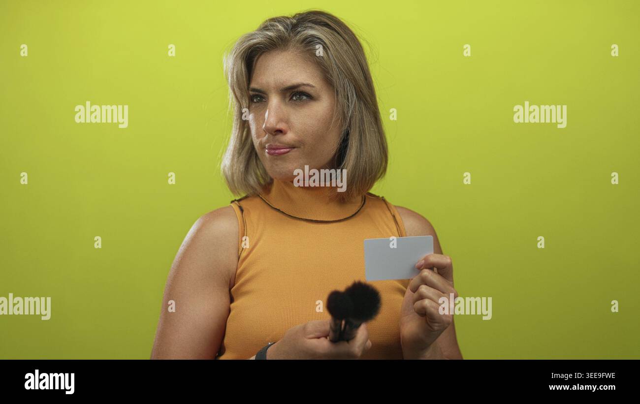 La giovane donna bionda esamina una spazzola per il trucco e una carta di plastica vuota con la fronte solcata in studio; indecisione. Foto Stock