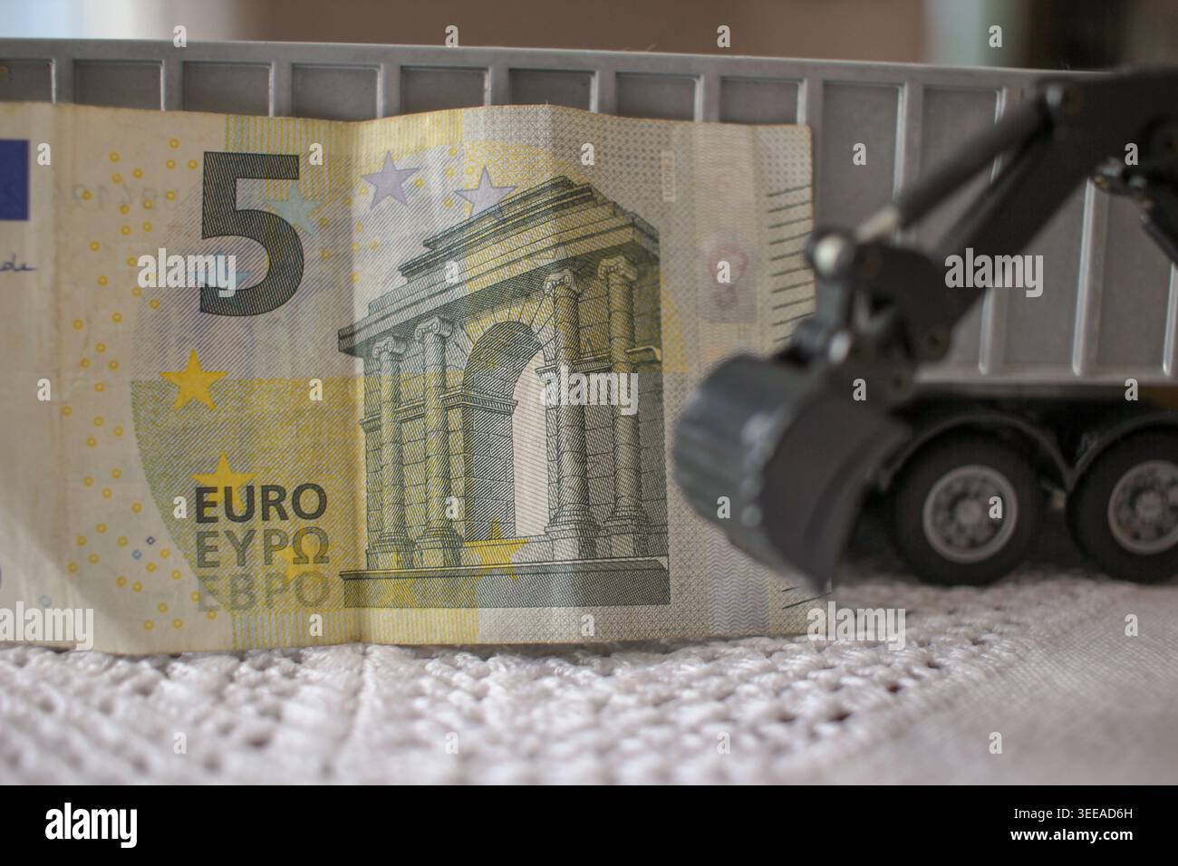 Una benna miniaturizzata per escavatore, concentrata di fronte a una banconota da 5 Euro, simboleggia i costi di lavori pubblici, progetti di ingegneria civile e sviluppo urbano Foto Stock