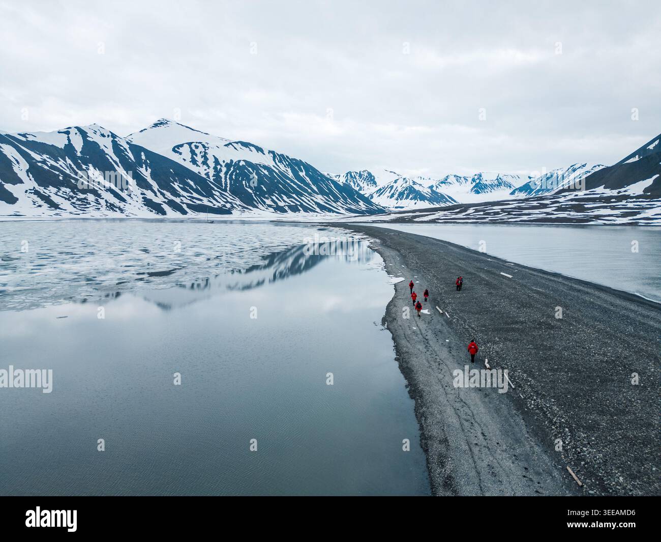 Svalbard e Jan Mayen - 2 luglio 2025: Veduta aerea delle persone in giacche rosse che camminano lungo uno stretto sputo di ghiaia tra le acque ghiacciate e la bocca innevata Foto Stock