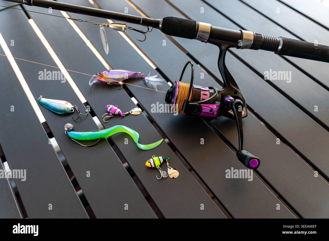 Lure da pesca assortite e canne posate su un tavolo con stecche scure, verme tenero verde brillante, Hardbait perlescente, maschera per perline, bobina di filatura viola con oro Foto Stock