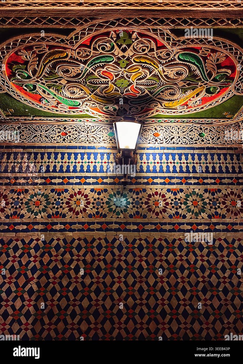 Piastrelle ornate in ceramica e soffitto intagliato illuminati da una lampada all'interno dello snack rack Café Riad nella vecchia Medina, 8 agosto 2025, Fez, Marocco Foto Stock