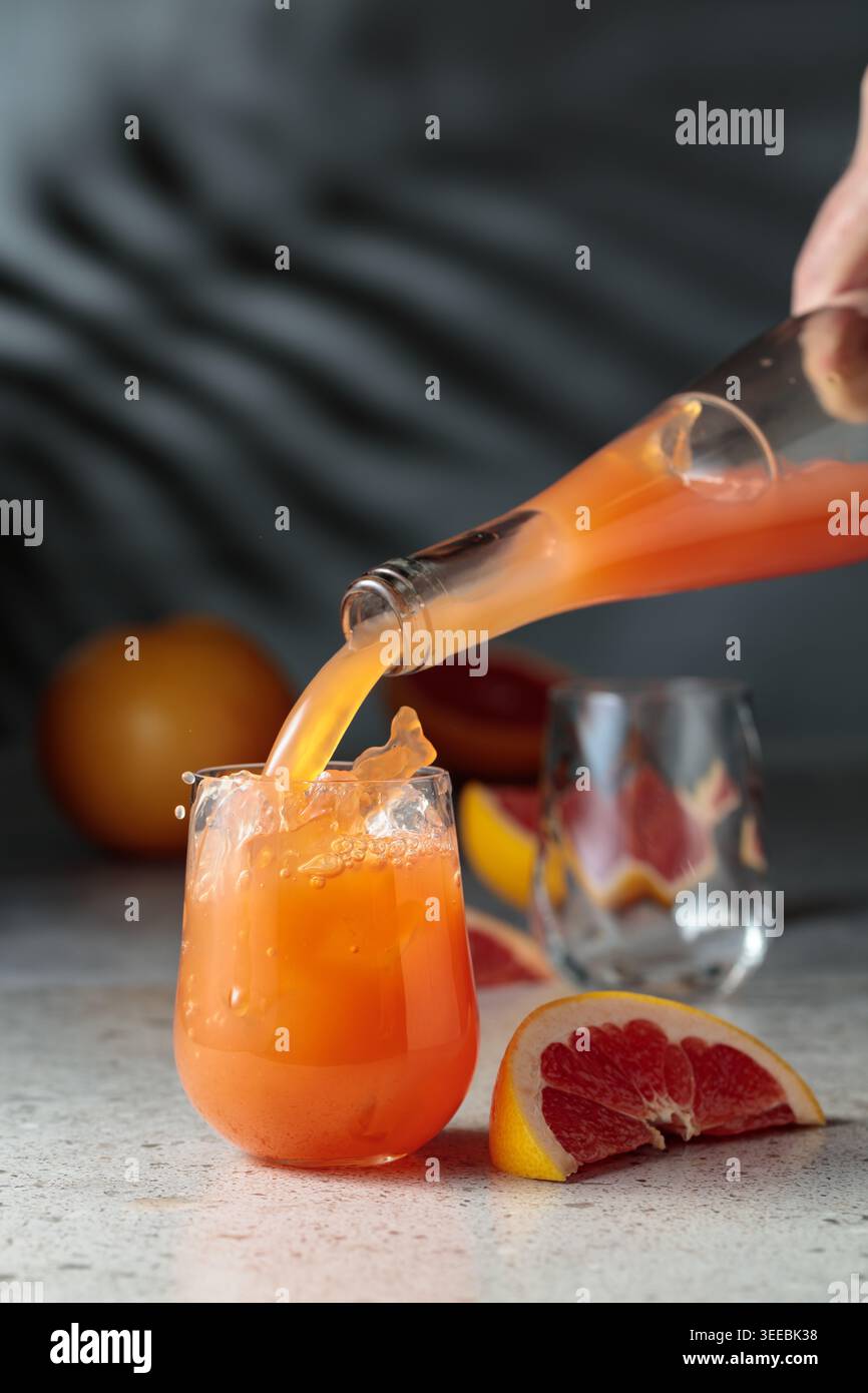 Il succo di pompelmo viene versato in un bicchiere con ghiaccio. Bere in un bicchiere congelato e frutta fresca su un tavolo di pietra. Sullo sfondo, le ombre del palmo leav Foto Stock