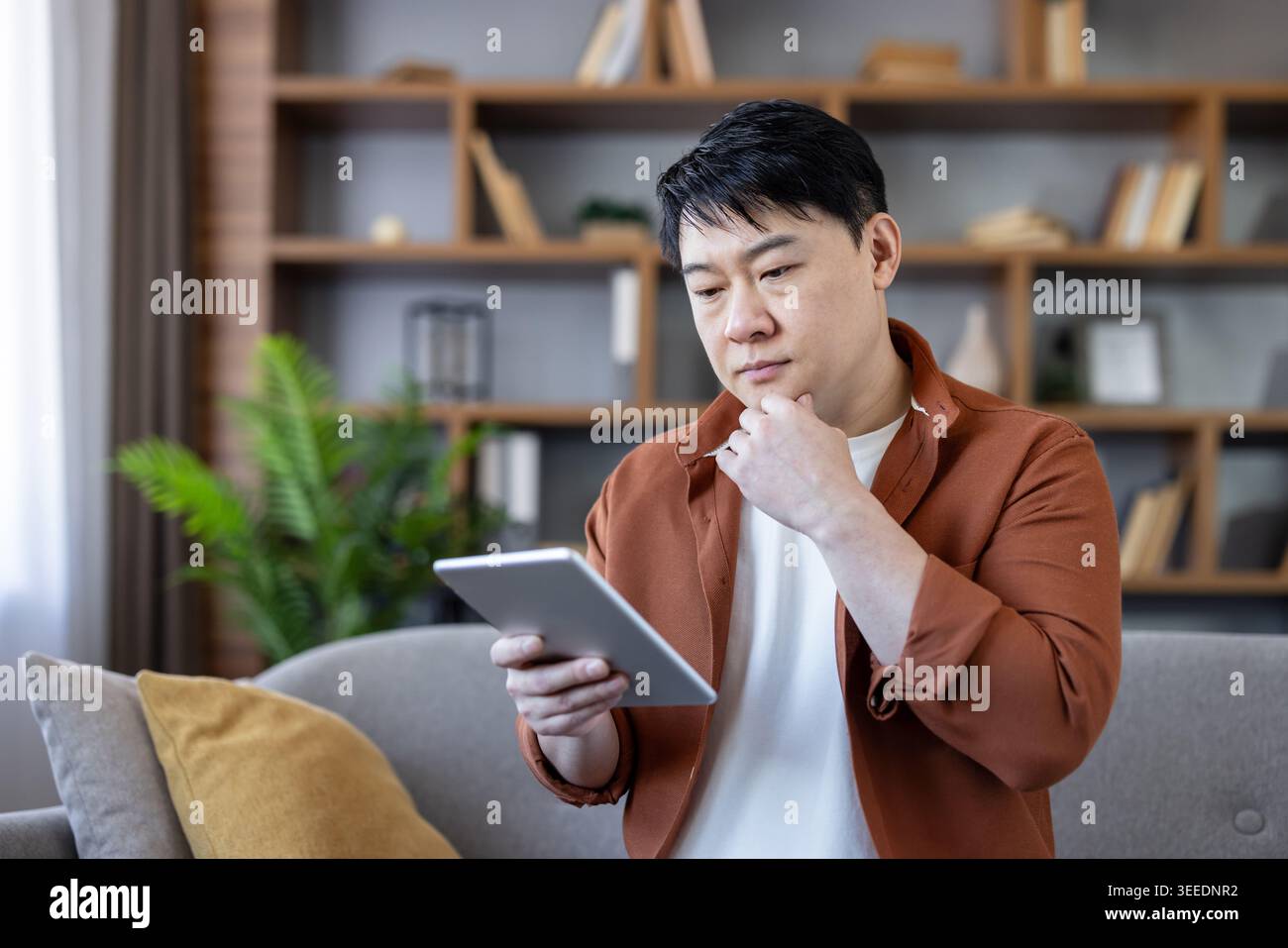 Uomo asiatico seduto su un divano che si concentra profondamente mentre legge un e-book su un tablet digitale, tenendo il mento, con una libreria e una pianta sullo sfondo Foto Stock