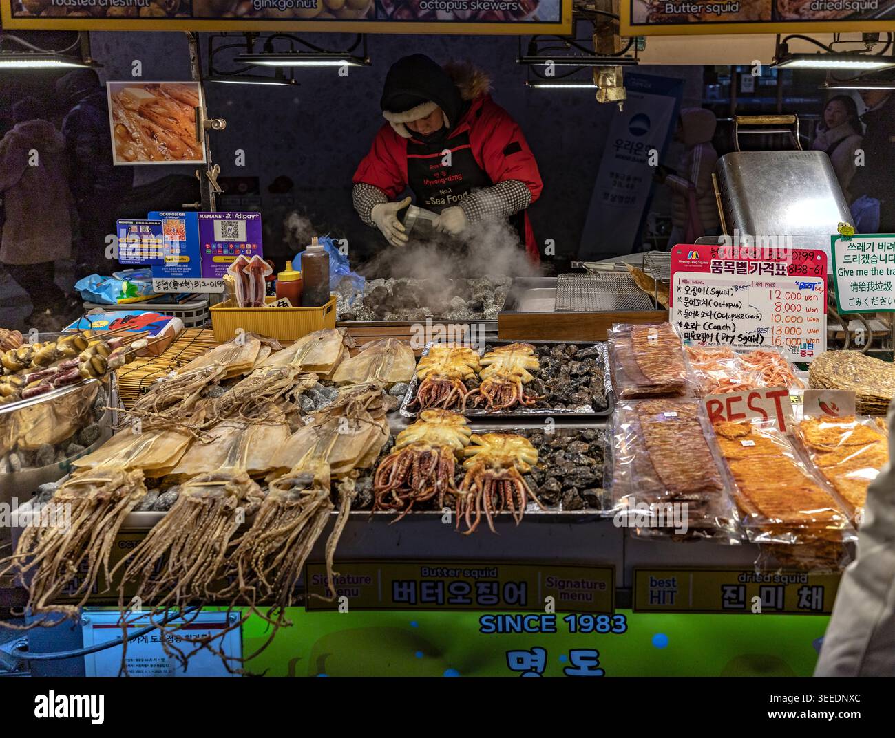 Venditore di bancarelle di frutti di mare che griglia calamari al burro al mercato notturno di Myeongdong a Seoul, Corea del Sud, con polpi, conchiglie e calamari essiccati in vendita Foto Stock