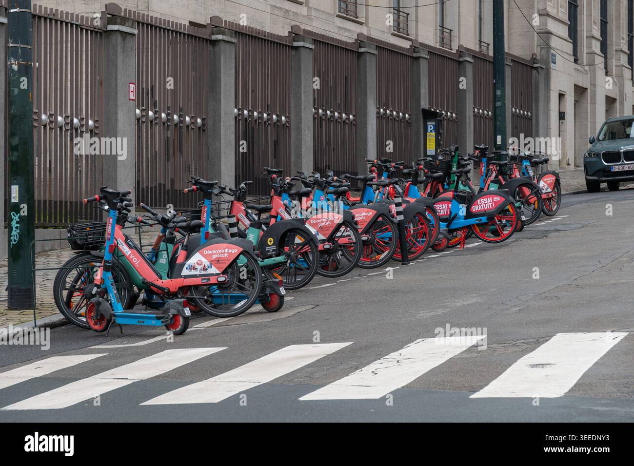 Bruxelles, Belgio - 2 aprile 2026: Linee condivise di scooter elettrici, strada cittadina vicino all'incrocio zebra, recinzione metallica, furgone parcheggiato, segnaletica stradale, urbano Foto Stock