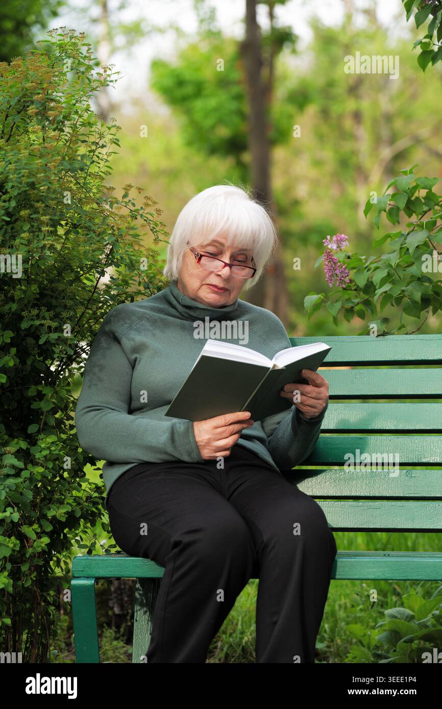Donna anziana in occhiali, che ama la letteratura e riposa all'aperto in un giardino primaverile. Donna in pensione che legge un romanzo su una panchina di legno nel parco, pace Foto Stock