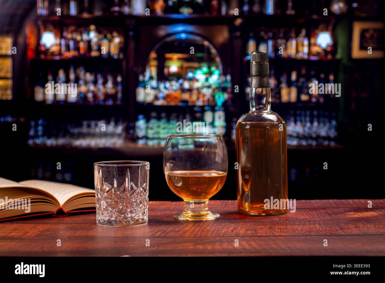 Libro aperto, bicchiere di cristallo, snifter di whisky e bottiglia su un bar di legno, con scaffali da pub irlandesi sfocati e calda atmosfera serale. Foto Stock