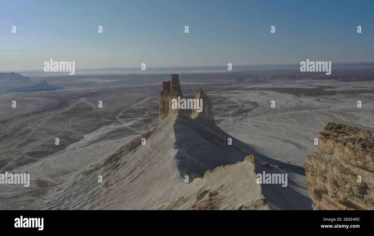 Una prospettiva aerea cattura le torreggianti formazioni rocciose che emergono drammaticamente da un vasto paesaggio desertico arido sotto un cielo azzurro limpido con sole tenue Foto Stock
