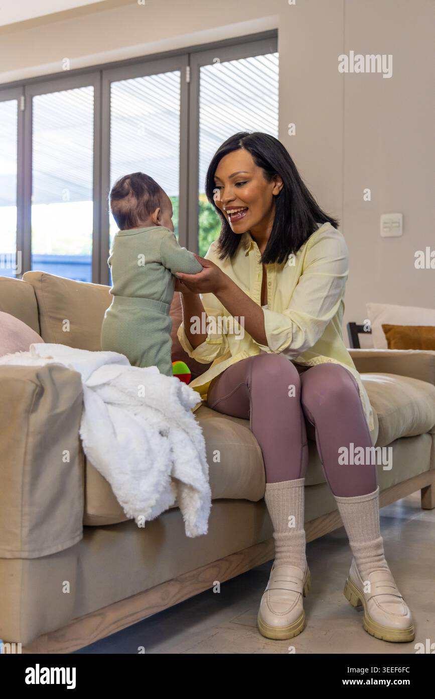 Madre e figlia afroamericana sedute su un divano beige tenendosi le mani con una camicia gialla Foto Stock