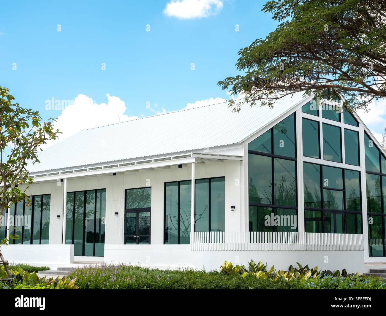 Moderna casa in vetro dall'architettura minimalista e grandi finestre, circondata da un giardino verde sotto il cielo blu. Design contemporaneo per la casa immobiliare Foto Stock