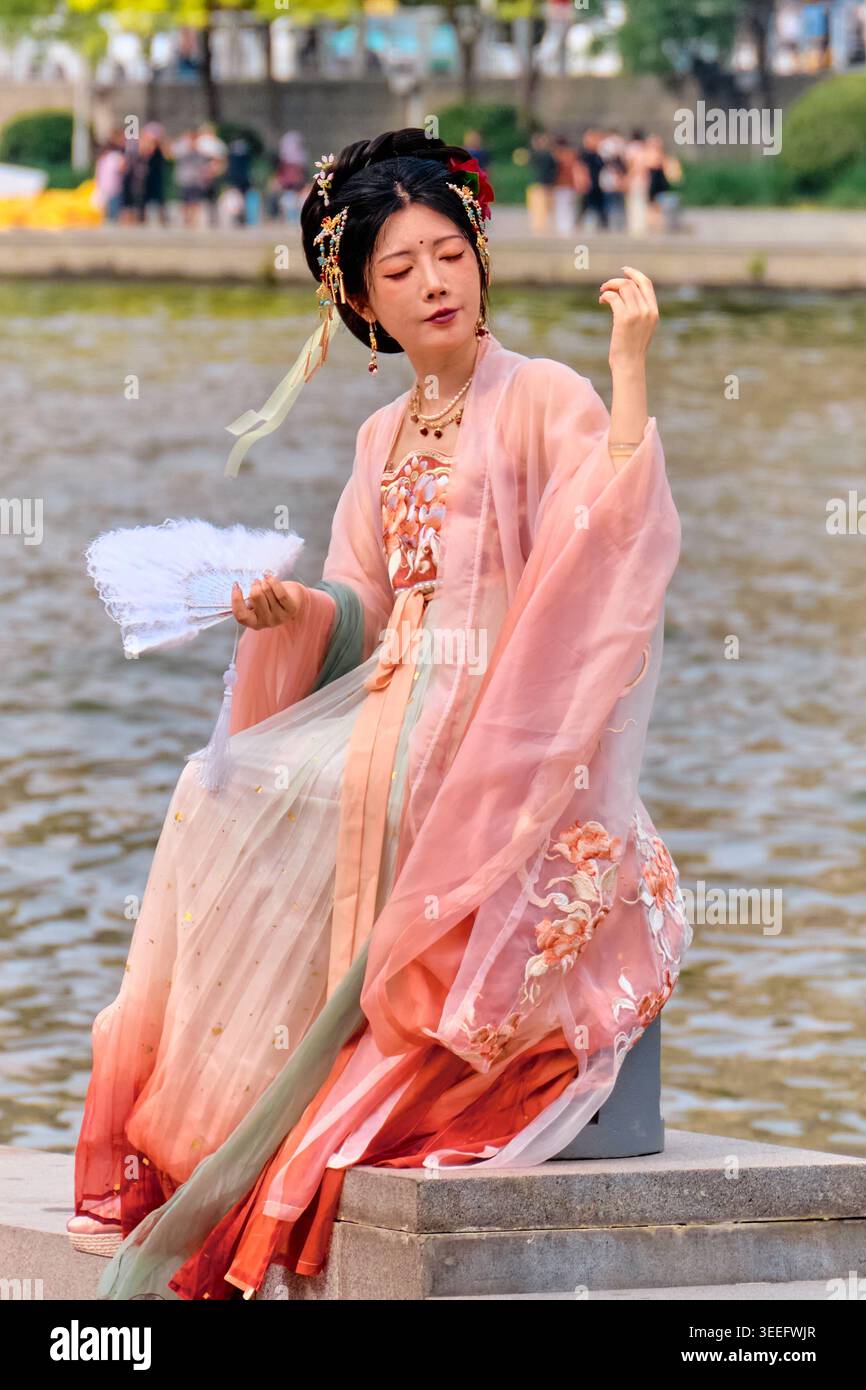 PECHINO, CINA - 01 giu 2025: Donna in hanfu tradizionale cinese seduta sulla panchina sul lungomare con ventilatore di piume bianco. Foto di alta qualità Foto Stock
