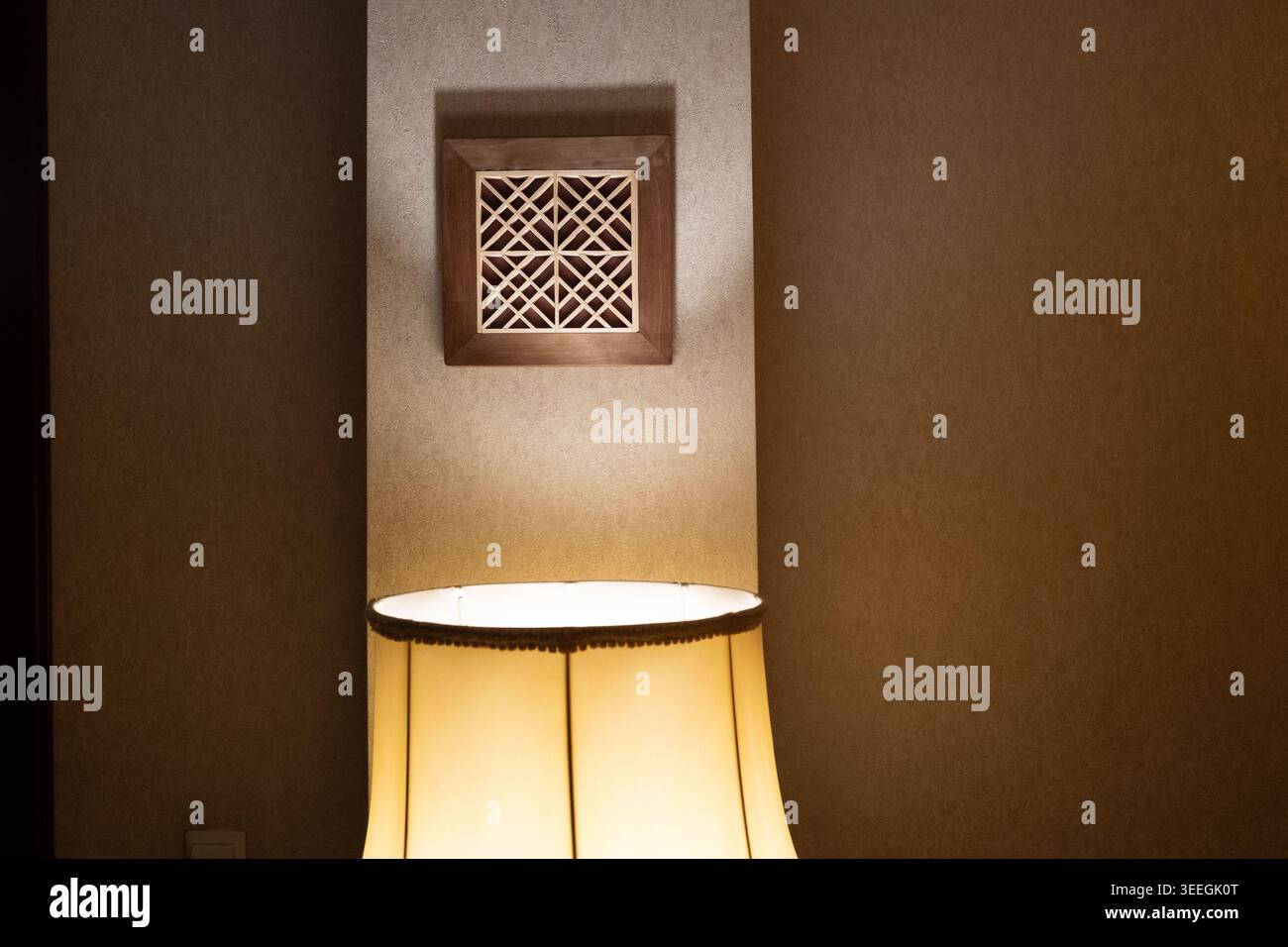 Una lampada da tavolo calda e luminosa diffonde una luce soffusa su una parete testurizzata sotto un pannello decorativo in legno, creando un'atmosfera intima e accogliente. Foto Stock