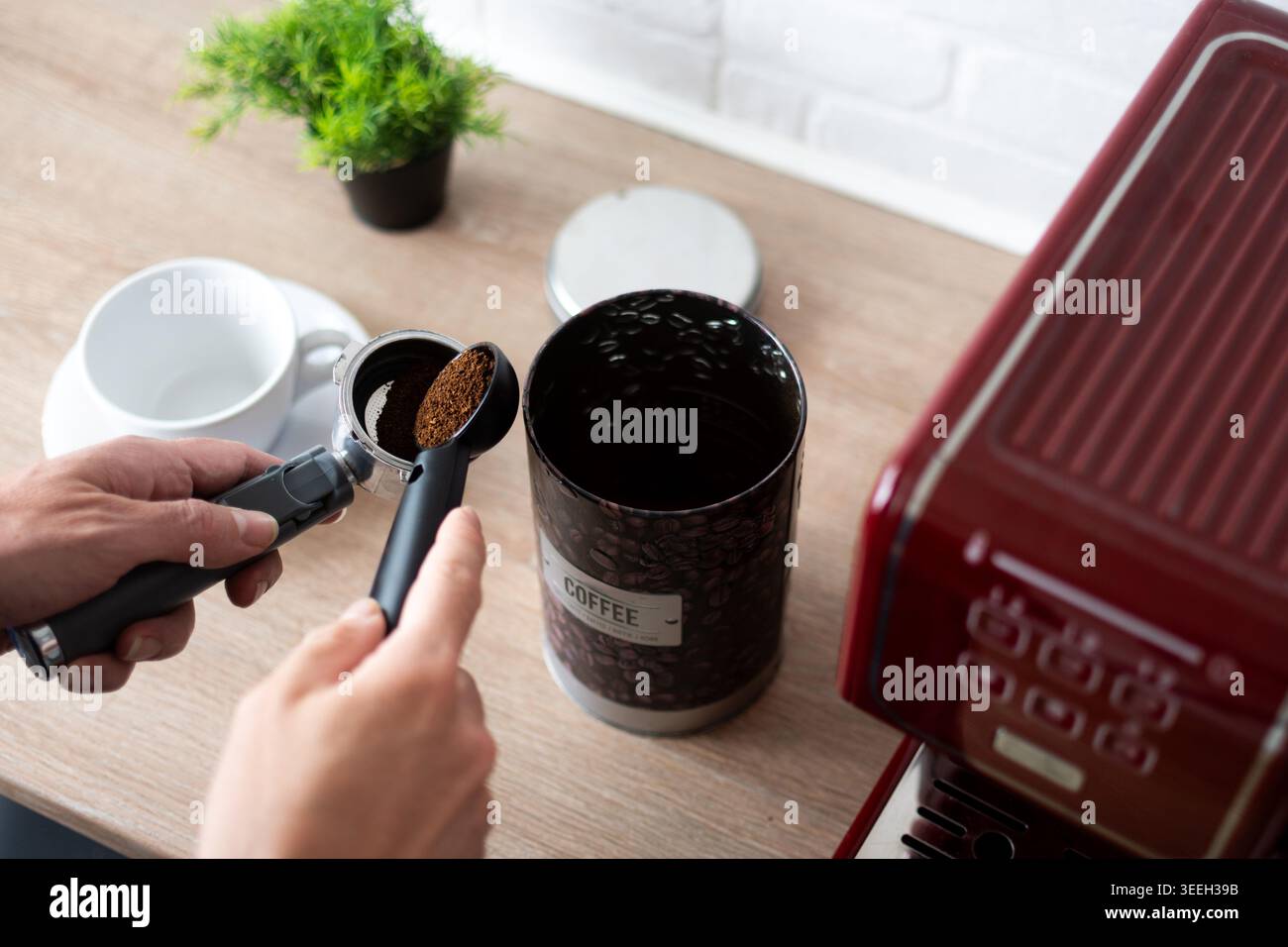 Primo piano di mani che riempiono il portafiltro con caffè macinato accanto a una macchina da caffè rossa e al contenitore del caffè su un piano di lavoro della cucina. Caffè a casa pr Foto Stock
