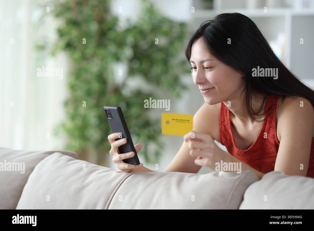 Donna asiatica soddisfatta che paga online con carta di credito e cellulare a casa Foto Stock