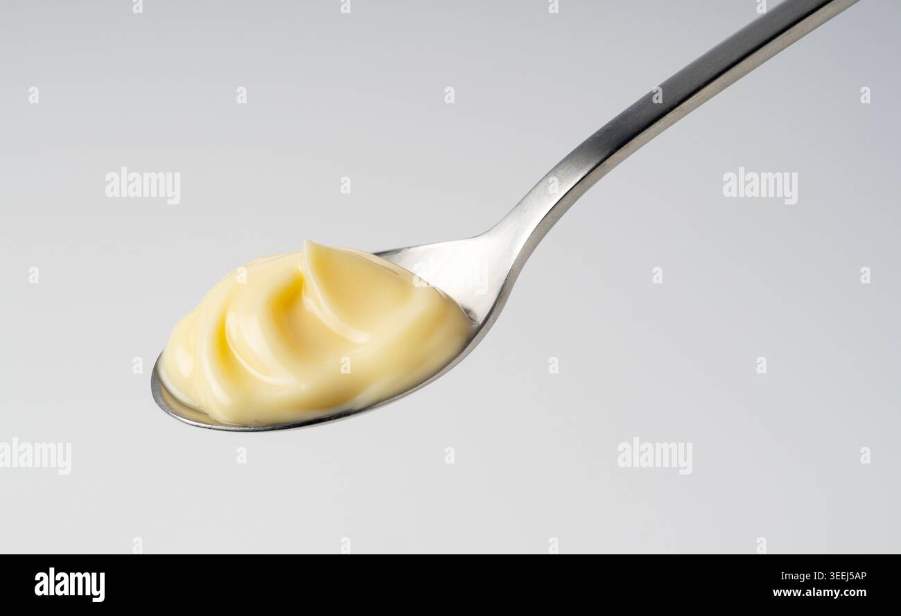 Maionese cremosa con elegante vortice su un cucchiaio in acciaio inossidabile, condimento in salsa gialla su sfondo grigio chiaro Foto Stock