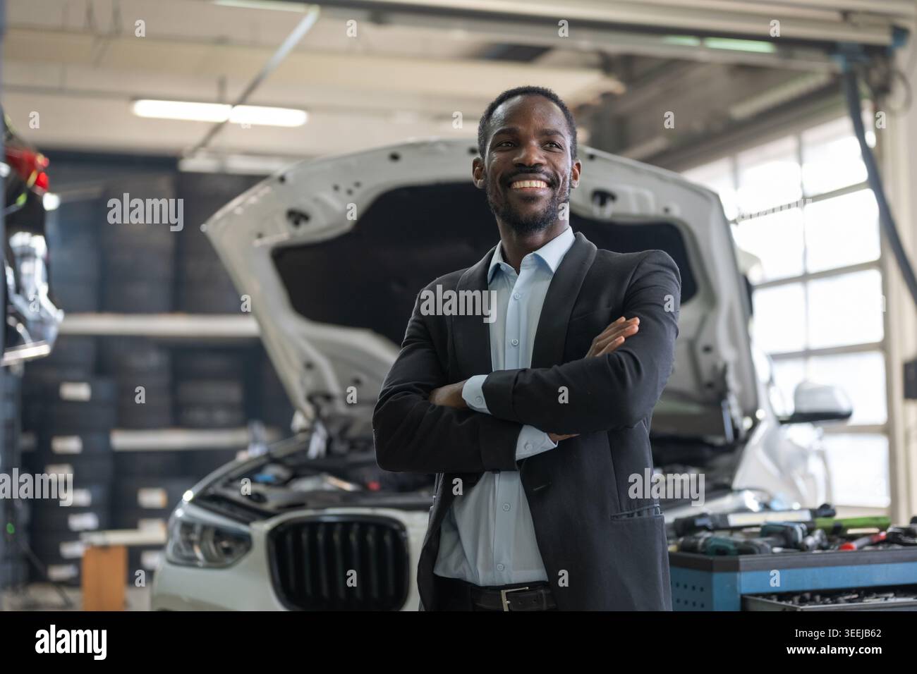 Un uomo d'affari sorride mentre supervisiona l'assistenza in un'officina di riparazione auto. Si trova di fronte a un veicolo di lusso con il cofano aperto. Foto Stock
