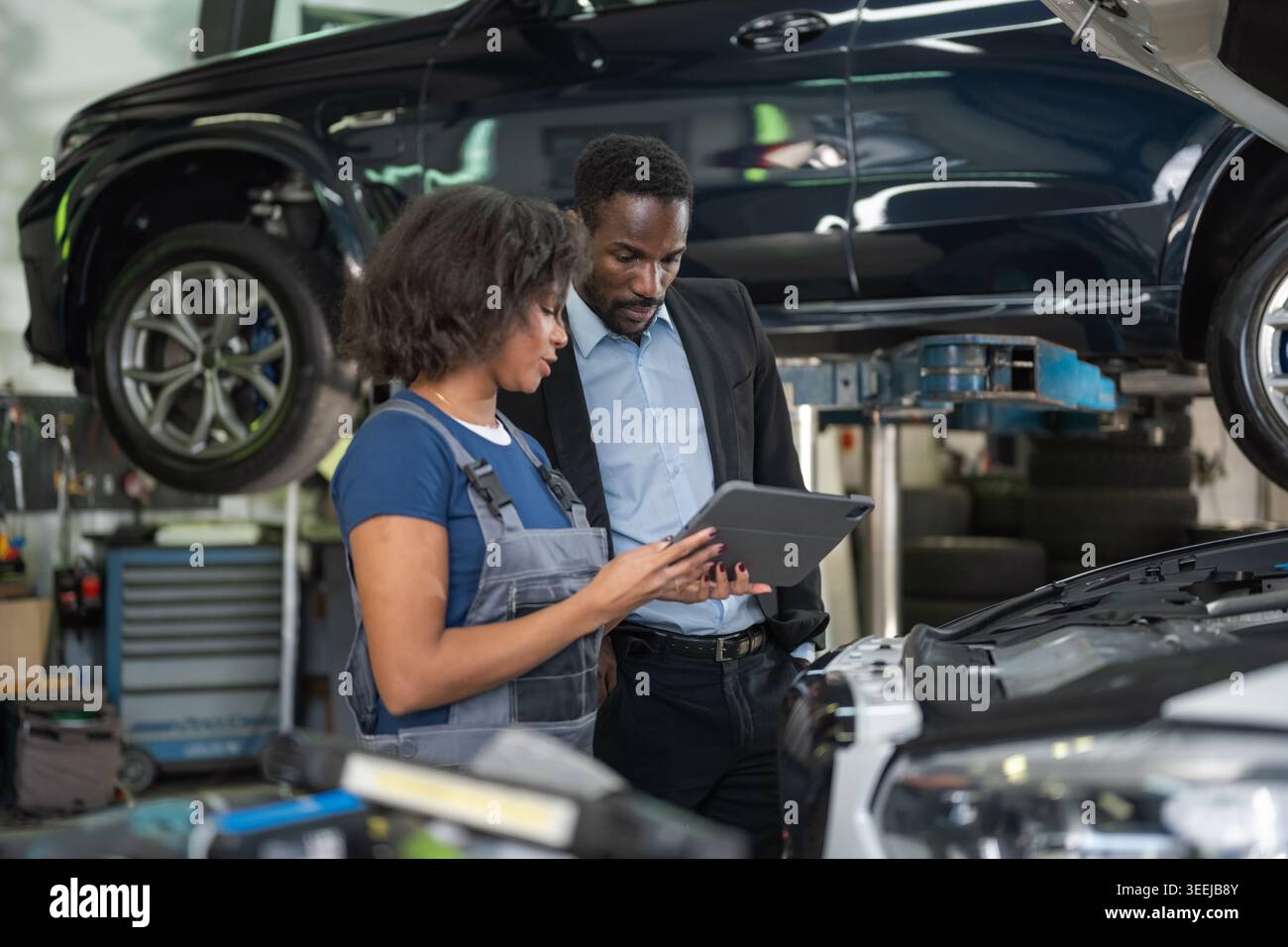 Meccanico donna che indica le parti del motore mentre si è in possesso di un tablet che visualizza i dati diagnostici, spiegando la riparazione a un cliente maschio. Foto Stock