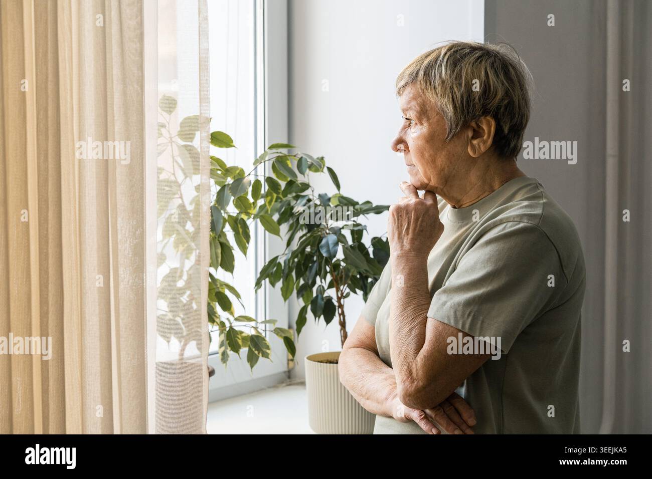 Donna anziana in piedi davanti a una finestra, che guarda all'esterno con un'espressione pensiva, che pensa alla solitudine. Sta riflettendo sulla solitudine e sull'invecchiamento Foto Stock