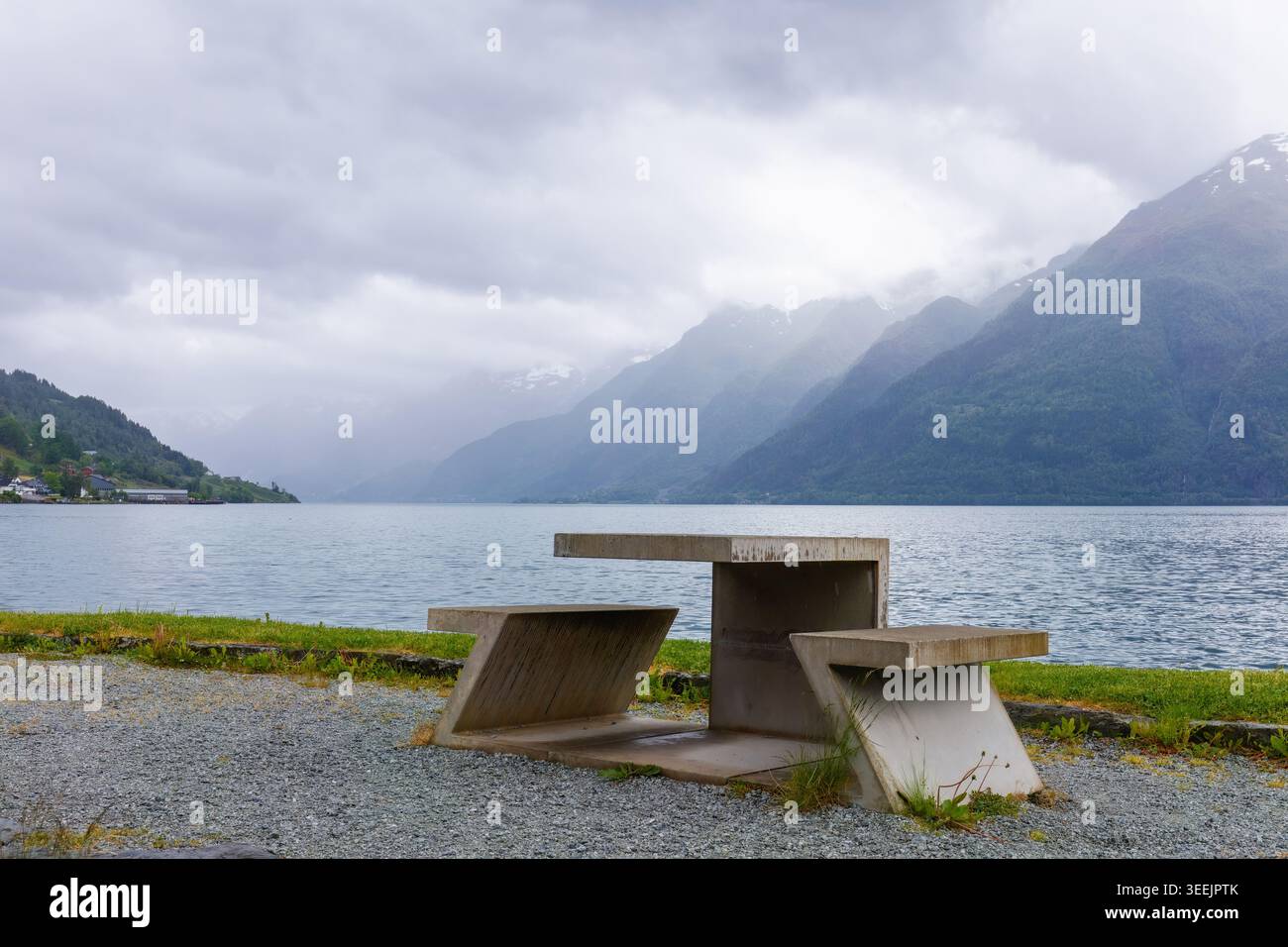 Fuga tranquilla dall'Hardangerfjord con tavolo in cemento che si affaccia sulle acque calme e montagne stratificate con nuvole di pioggia nella Norvegia occidentale. Viaggi contemplativi, S Foto Stock
