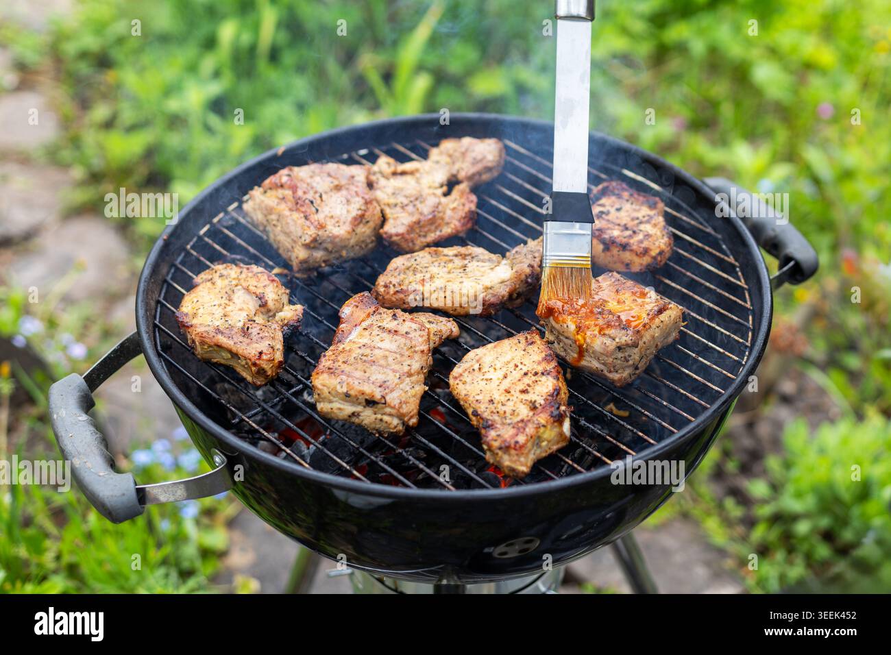 Cottura di carne alla griglia su barbecue a carbone. I pezzi di carne conditi cuocono su una griglia a carbone affumicato mentre la salsa viene spennellata in un cortile esterno Foto Stock