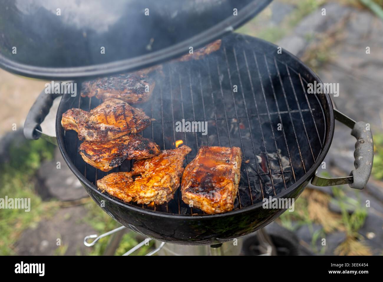 Carne barbecue affumicata su griglia a carbone. I pezzi di carne glassati si mescolano su un barbecue a carbone affumicato con fiamme e aprono il coperchio durante la cottura all'aperto. Foto Stock