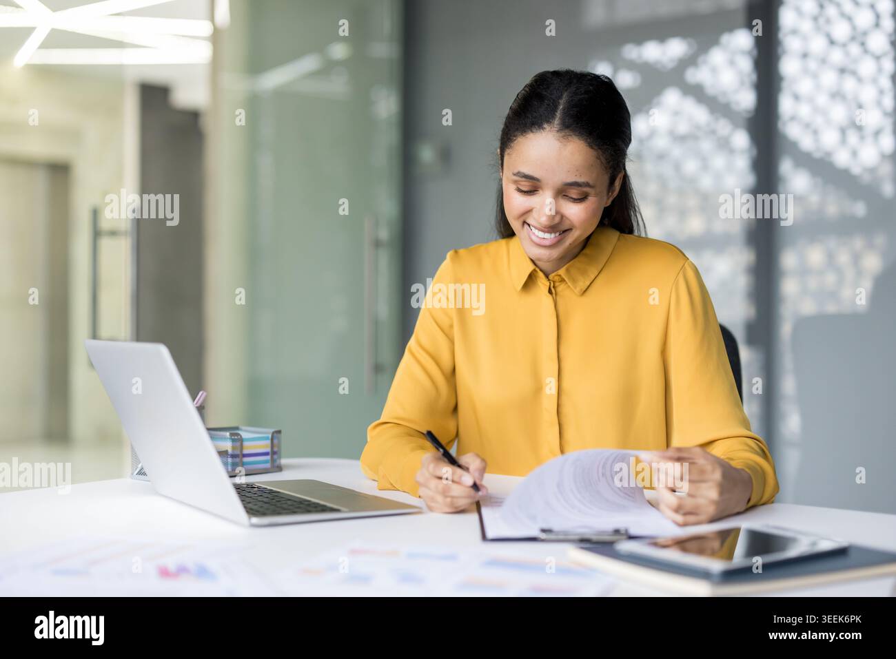 Giovane donna d'affari mista sorridente mentre esamina e firma importanti documenti contrattuali, gestisce i documenti cartacei e le responsabilità presso una scrivania in un ambiente d'ufficio moderno Foto Stock