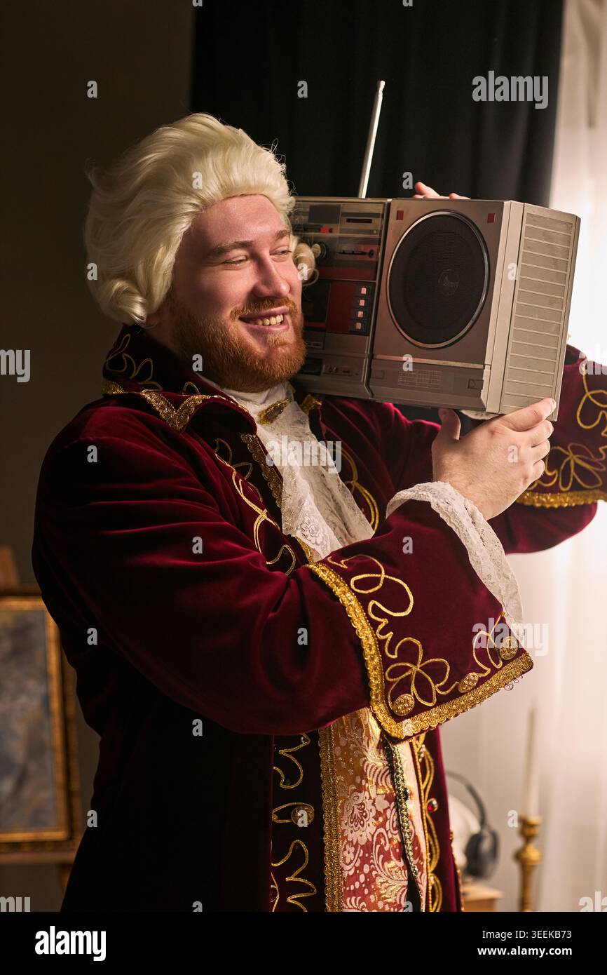 Ripresa verticale di un giovane uomo in costume storico che regge il boombox sulla spalla, sorride e posa con un concetto di musica retrò. Ideale per l'umorismo, la nostalgia, il marketing dell'intrattenimento Foto Stock