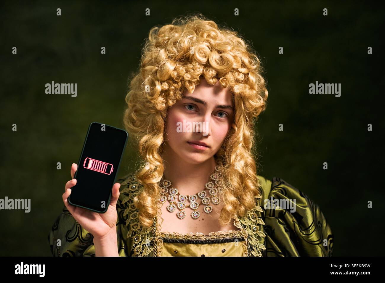 Ritratto di una giovane donna che tiene in mano uno smartphone, che mostra l'icona della batteria scarica, che illustra la mancanza di alimentazione e la dipendenza digitale. Utile per l'affaticamento tecnico, la ricarica e gli annunci di dispositivi Foto Stock