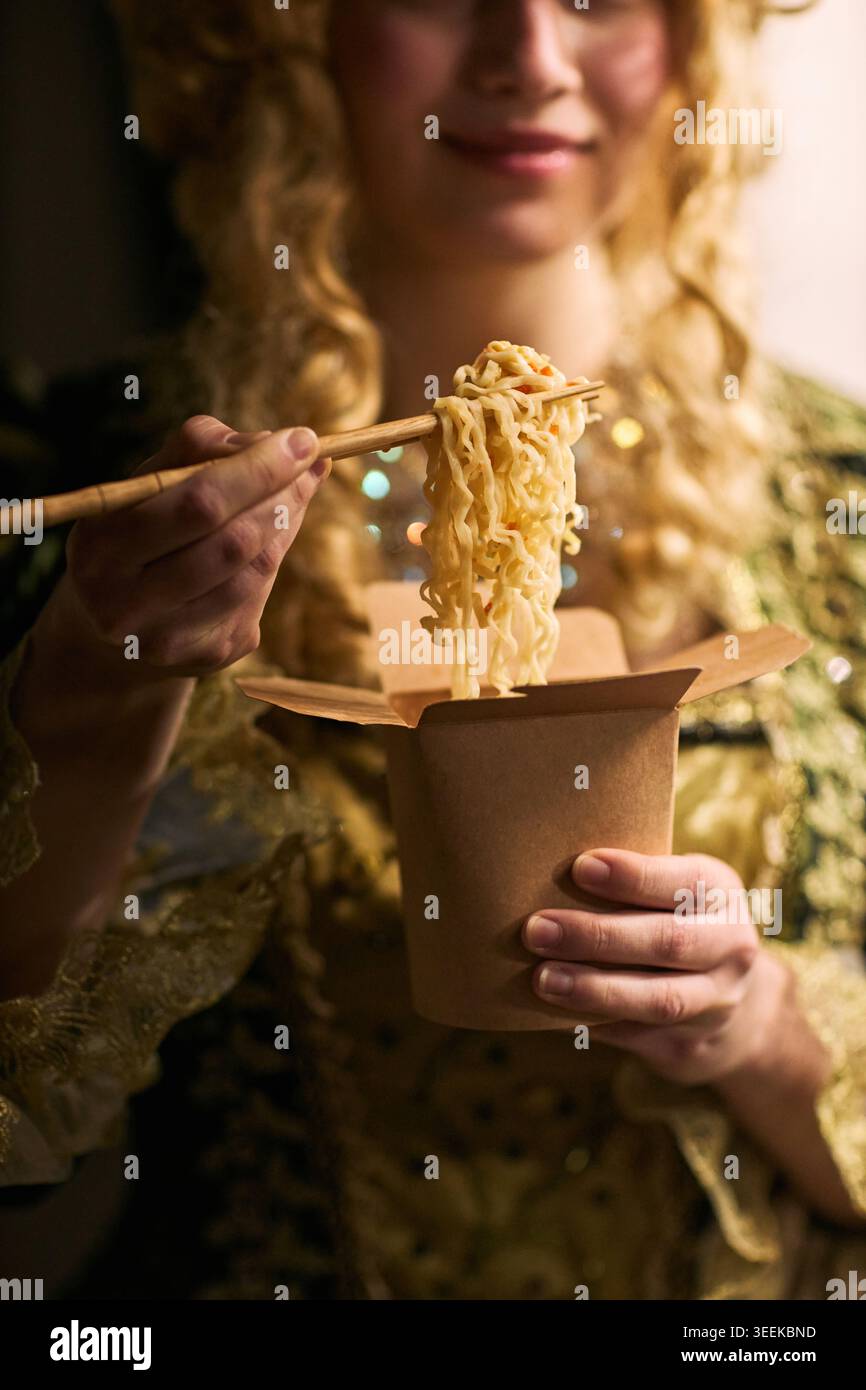Immagine verticale di una giovane donna che mangia spaghetti dalla scatola da asporto con bacchette, mostrando un concetto pratico di pasto veloce. Utile per la consegna di cibo, asporto, marketing rapido per il pranzo Foto Stock