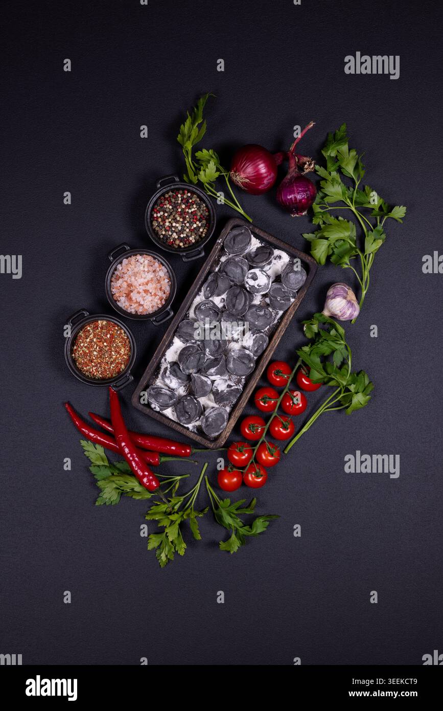 Pelmeni neri con impasto scuro e ripieno cremoso disposti in un vassoio tra pomodori ciliegini, cipolla rossa, aglio, prezzemolo, peperoncino e ciotole per spezie Foto Stock