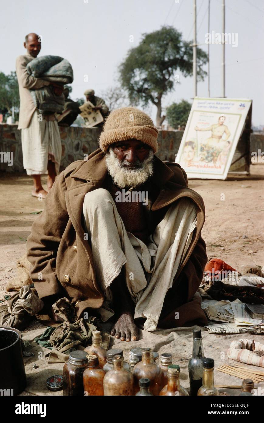 Uno scivolo da viaggio vintage a colori d'archivio dei primi anni '1970 che raffigura un anziano indiano che indossa un cappotto pesante e una copertura per la testa, seduto all'aperto a terra in un'area del mercato di nuova Delhi. Foto Stock