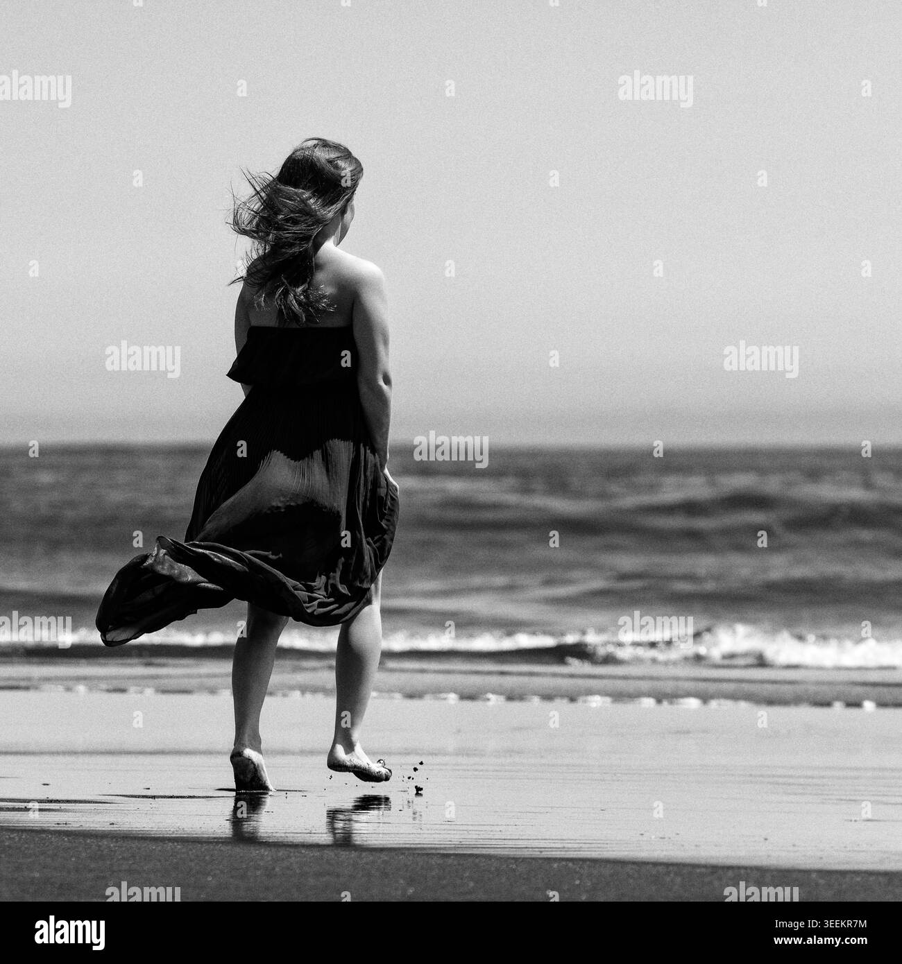 Donna in piedi sulla spiaggia vicino all'oceano. Donna in abito che cammina sulla riva del mare. Fotografia in bianco e nero con disturbo retrò ed effetto granulosità analogico. Cinema Foto Stock